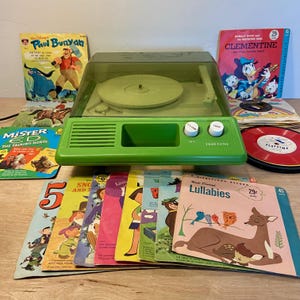 May include: A vintage green Truetone record player with a clear lid, surrounded by colorful children's records. Album covers feature characters like Donald Duck and Paul Bunyan. The record player has volume and tone controls.