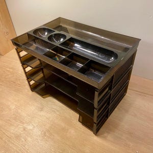 May include: A dark brown translucent plastic desk organizer featuring a tiered design with three open shelves and a top tray. The top tray includes two circular compartments and a long rectangular section for storing small office supplies.