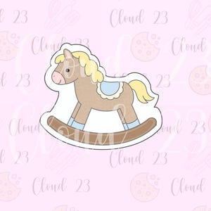 May include: A cookie cutter in the shape of a rocking horse. The horse is brown with a yellow mane and tail, a pink nose, and a blue saddle. The background is light pink with the words "Cloud 23" repeated.