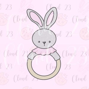 May include: A gray bunny-shaped cookie cutter with pink inner ears and a pink ruffle collar. The bunny is holding a tan ring. The background is light pink with the text "Cloud 23" repeated.