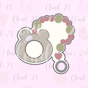 May include: A cookie cutter in the shape of a bear with a beaded necklace. The bear is light brown with a white center and a silver outline. The necklace has green and red beads. The background is pink with the text "Cloud 23".