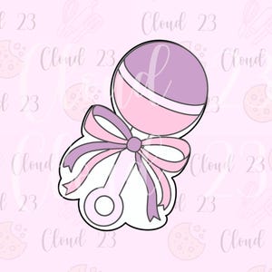 May include: A cookie cutter in the shape of a baby rattle. The rattle is light pink and purple with a large bow. The text "Cloud 23" is repeated across the background.