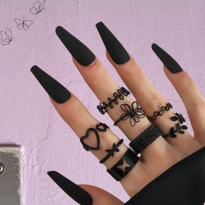 Könnte beinhalten: Eine Hand mit mehreren schwarzen Ringen mit verschiedenen Designs, darunter Herzen, Schmetterlinge und Blumenmuster. Die Nägel sind schwarz lackiert. Der Hintergrund ist helllila.