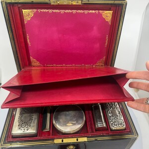 May include: An open antique vanity case with a black exterior, red interior, and gold trim. The case contains several silver-toned accessories. The lid is open, revealing a vibrant pink lining and gold decorative accents.