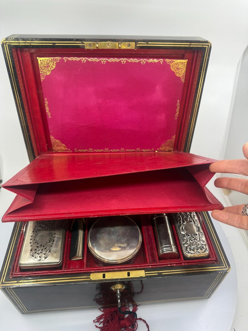 May include: An open antique vanity case with a black exterior, red interior, and gold trim. The case contains several silver-toned accessories. The lid is open, revealing a vibrant pink lining and gold decorative accents.