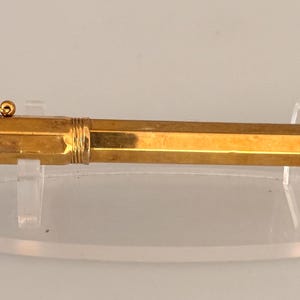 May include: A gold-colored fountain pen with a hexagonal barrel and cap. The pen features a thin, straight pocket clip with a small spherical finial at the end. The cap and barrel are connected by a threaded section, and the end of the barrel is threaded.