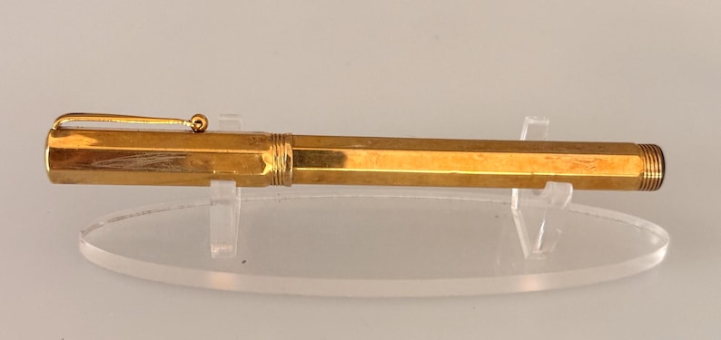 May include: A gold-colored fountain pen with a hexagonal barrel and cap. The pen features a thin, straight pocket clip with a small spherical finial at the end. The cap and barrel are connected by a threaded section, and the end of the barrel is threaded.