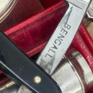 May include: Close-up of a vintage shaving kit with a black handled straight razor, a silver-colored blade with the word "BENGALL" etched on it, and other grooming tools. The kit is housed in a red velvet-lined case.