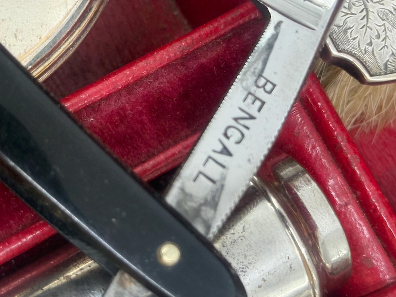 May include: Close-up of a vintage shaving kit with a black handled straight razor, a silver-colored blade with the word "BENGALL" etched on it, and other grooming tools. The kit is housed in a red velvet-lined case.