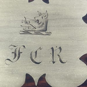 May include: Close-up of a decorative metal plate with a boar's head crest and the initials FCR in elegant script. The plate is set against a dark wood background, showcasing a vintage aesthetic.