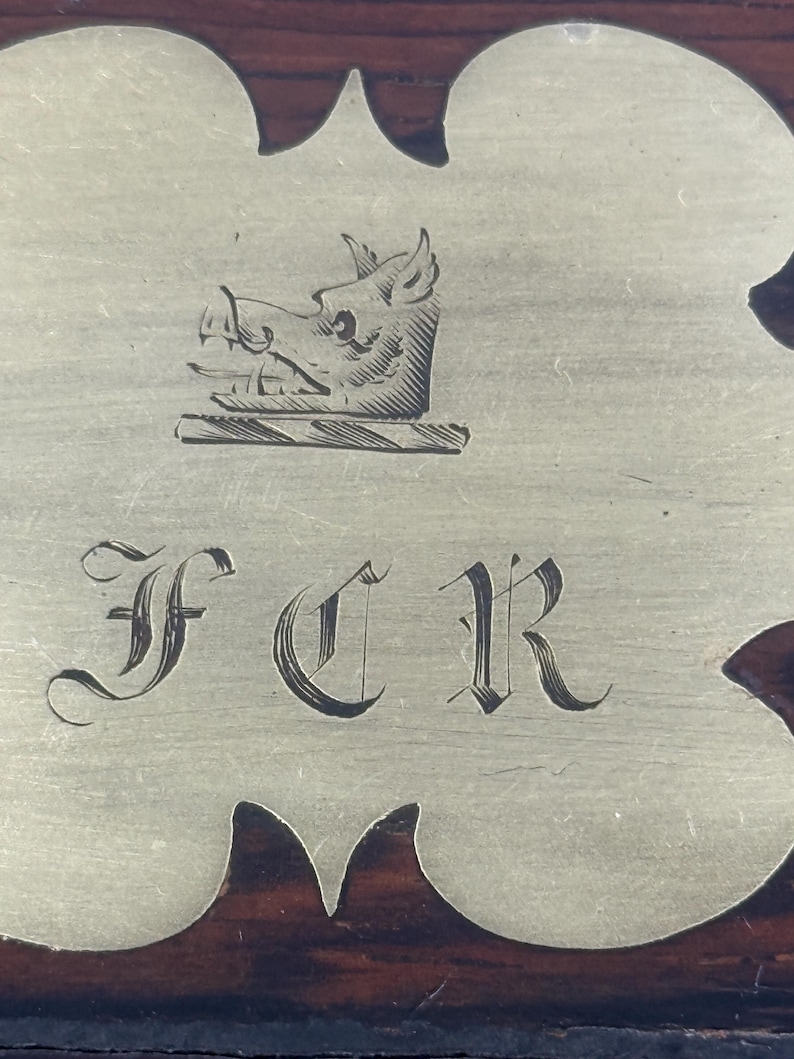 May include: Close-up of a decorative metal plate with a boar's head crest and the initials FCR in elegant script. The plate is set against a dark wood background, showcasing a vintage aesthetic.
