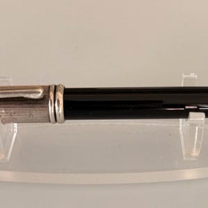 Puede incluir: Una pluma estilográfica con cuerpo negro y capuchón plateado con acabado texturizado y acanalado. La pluma incluye un clip de bolsillo plateado y un tapón final a juego, presentada horizontalmente sobre un soporte de exhibición de acrílico transparente.