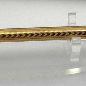 Puede incluir: Un portaminas de tono dorado con un cuerpo cilíndrico delgado y un intrincado patrón de espiga grabado a lo largo de su longitud. El lápiz presenta una tapa decorativa redondeada en el extremo y una punta fina con una mina visible.