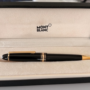 May include: A black Montblanc ballpoint pen with gold-colored accents, presented in an open black box with a cream-colored interior. The Montblanc logo is visible on the box's interior.