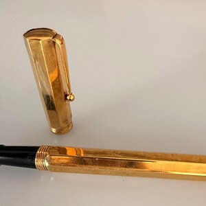 May include: A gold-toned fountain pen with a hexagonal barrel and a matching removable cap. The pen features a black grip section and a metallic gold nib. The cap includes a sturdy pocket clip and a rounded top, showcasing a polished finish.