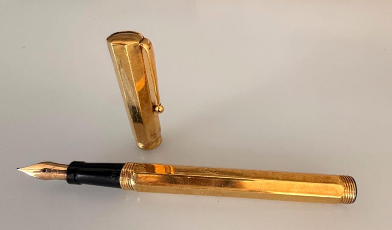 May include: A gold-toned fountain pen with a hexagonal barrel and a matching removable cap. The pen features a black grip section and a metallic gold nib. The cap includes a sturdy pocket clip and a rounded top, showcasing a polished finish.