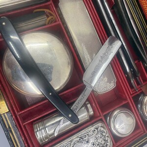 May include: An antique shaving kit with a black handled straight razor, a silver-toned mirror, and various grooming tools. The kit is housed in a red-lined case with multiple compartments, suggesting a vintage or collectible item.