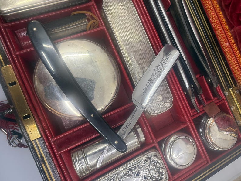 May include: An antique shaving kit with a black handled straight razor, a silver-toned mirror, and various grooming tools. The kit is housed in a red-lined case with multiple compartments, suggesting a vintage or collectible item.