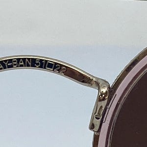 Puede incluir: Primer plano del brazo de metal plateado de unas gafas de sol redondas. El brazo est&aacute; grabado con el texto "RAY-BAN 51□22".