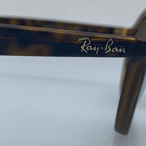 May include: Close-up of a pair of tortoiseshell Ray-Ban sunglasses. The sunglasses have a gold logo on the side.
