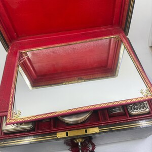 May include: Open red leather jewelry box with a mirrored lid and gold trim. The interior is red with compartments for small items. A key with a red tassel hangs from the lock.