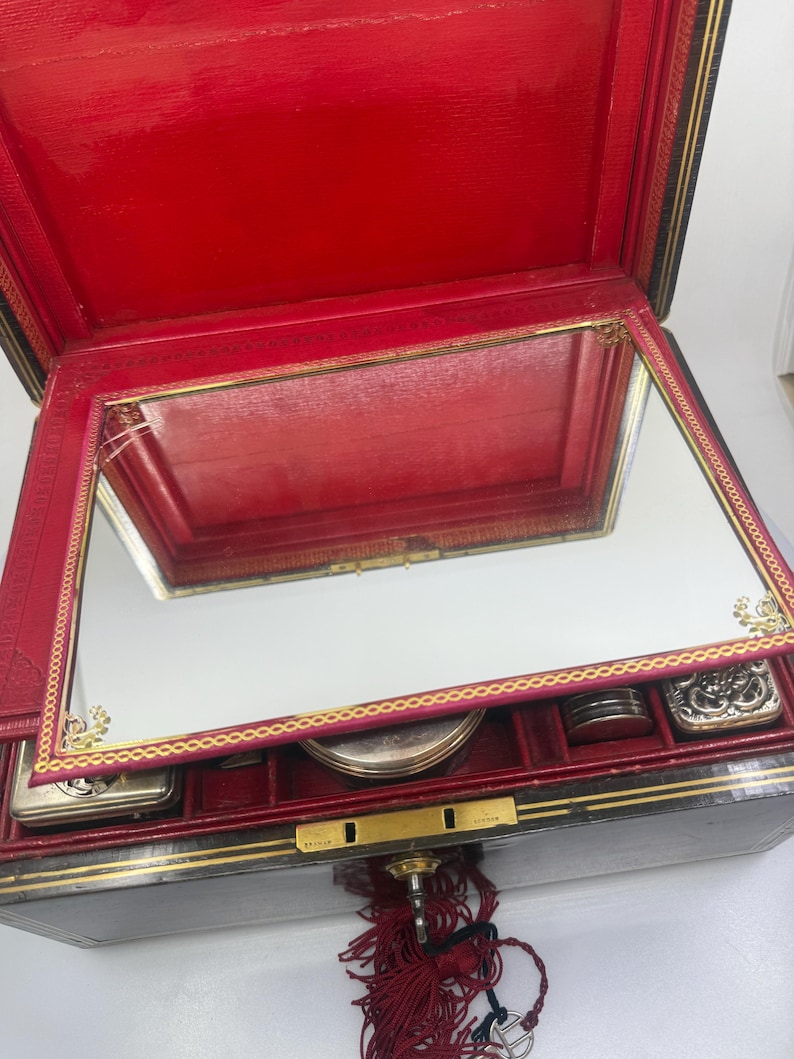 May include: Open red leather jewelry box with a mirrored lid and gold trim. The interior is red with compartments for small items. A key with a red tassel hangs from the lock.