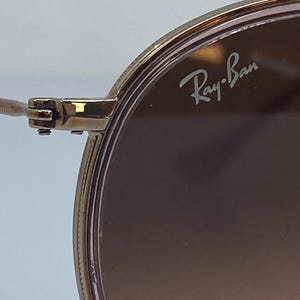 Puede incluir: Primer plano de unas gafas de sol con montura dorada y lentes de color rosa. El logotipo de Ray-Ban es visible en la lente derecha.