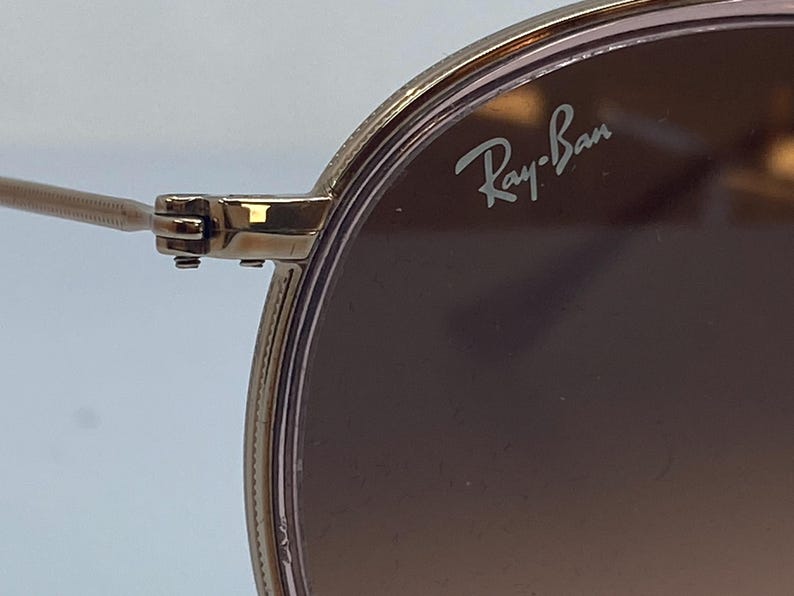 Puede incluir: Primer plano de unas gafas de sol con montura dorada y lentes de color rosa. El logotipo de Ray-Ban es visible en la lente derecha.