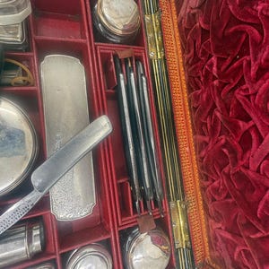 May include: An open antique grooming kit with a black-handled straight razor, silver-toned accessories, and various grooming tools. The interior is lined with red velvet, and the kit is divided into compartments.