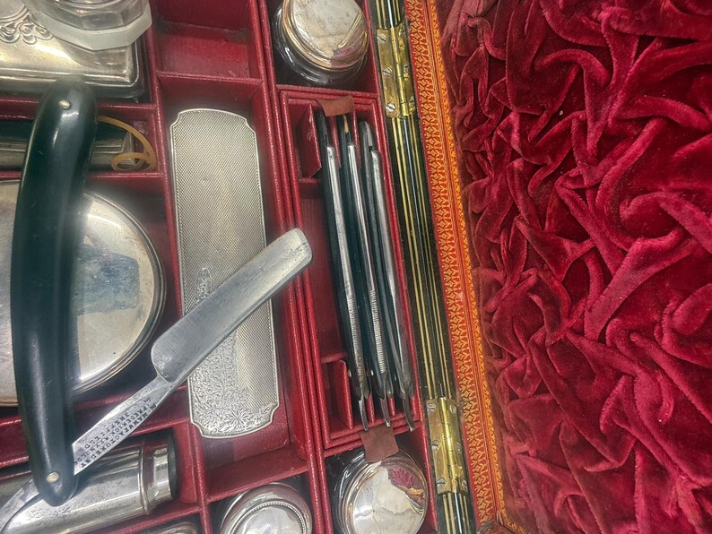 May include: An open antique grooming kit with a black-handled straight razor, silver-toned accessories, and various grooming tools. The interior is lined with red velvet, and the kit is divided into compartments.