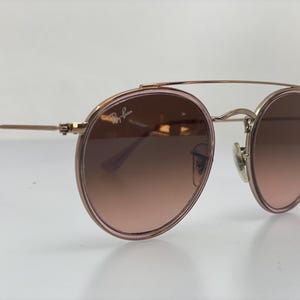 Puede incluir: Gafas de sol redondas de oro rosa con doble puente y lentes de color rosa degradado. El logotipo de Ray-Ban es visible en la lente derecha.