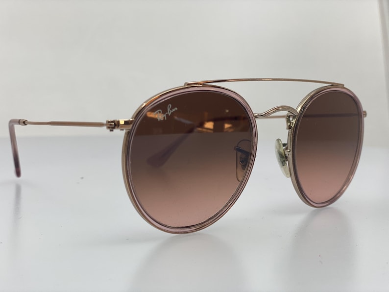 Puede incluir: Gafas de sol redondas de oro rosa con doble puente y lentes de color rosa degradado. El logotipo de Ray-Ban es visible en la lente derecha.