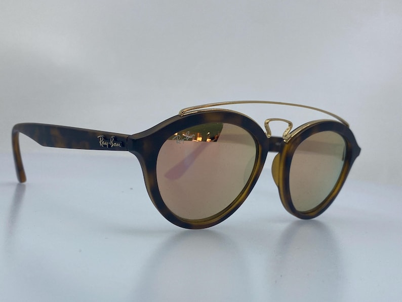 May include: Tortoise shell Ray-Ban sunglasses with gold accents and rose-colored lenses.