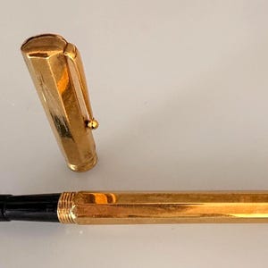 May include: A gold-toned fountain pen with a hexagonal barrel and a matching removable cap. The pen features a black grip section and a metallic gold nib. The cap includes a sturdy clip and a rounded top, showcasing a polished, reflective finish.