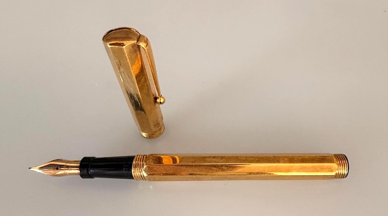 May include: A gold-toned fountain pen with a hexagonal barrel and a matching removable cap. The pen features a black grip section and a metallic gold nib. The cap includes a sturdy clip and a rounded top, showcasing a polished, reflective finish.