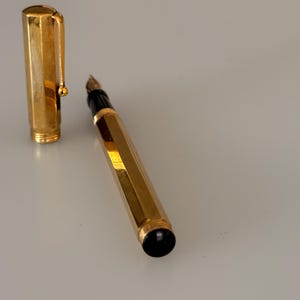 May include: A gold-toned fountain pen with a hexagonal barrel and a matching removable cap. The pen features a black grip section near the nib and a black circular end piece. The cap includes a gold-toned clip with a small spherical tip.