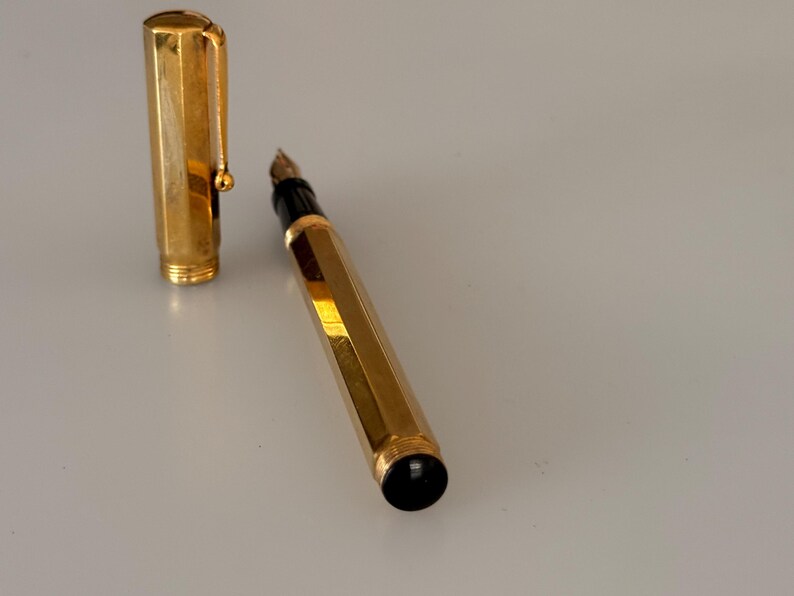 May include: A gold-toned fountain pen with a hexagonal barrel and a matching removable cap. The pen features a black grip section near the nib and a black circular end piece. The cap includes a gold-toned clip with a small spherical tip.