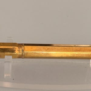 May include: A gold-colored fountain pen with a hexagonal barrel and a matching cap featuring a gold clip. The pen has a threaded section at the end of the barrel and is displayed horizontally on a clear stand, showcasing its metallic finish.