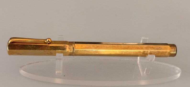 May include: A gold-colored fountain pen with a hexagonal barrel and a matching cap featuring a gold clip. The pen has a threaded section at the end of the barrel and is displayed horizontally on a clear stand, showcasing its metallic finish.