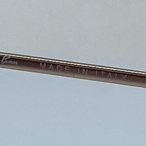 Puede incluir: Primer plano de una montura de metal plateada con el texto "Ray-Ban MADE IN ITALY" grabado en ella.
