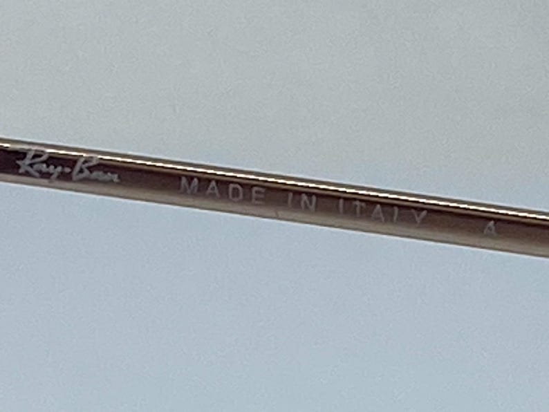 Puede incluir: Primer plano de una montura de metal plateada con el texto "Ray-Ban MADE IN ITALY" grabado en ella.