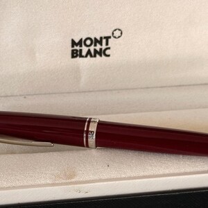 Könnte beinhalten: Ein bordeauxfarbener Montblanc-Stift mit silberfarbenen Akzenten liegt in einer weißen Schachtel. Der Stift hat ein schlankes Design mit einem silbernen Clip und einem silbernen Ring. Das Montblanc-Logo ist auf der Schachtel zu sehen.