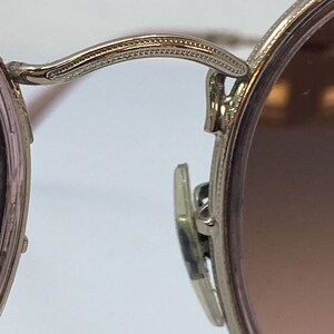Puede incluir: Primer plano del bisagra de unas gafas de sol redondas de color plateado. La bisagra tiene un dise&ntilde;o decorativo texturizado.