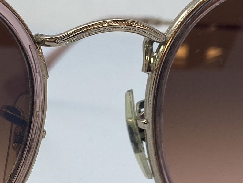 Puede incluir: Primer plano del bisagra de unas gafas de sol redondas de color plateado. La bisagra tiene un dise&ntilde;o decorativo texturizado.