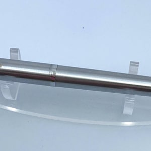 Op de afbeelding: Een zilverkleurige pen met een goudkleurige clip, tentoongesteld op een heldere acrylstandaard. De pen heeft een strak, modern ontwerp en de gouden clip voegt een vleugje elegantie toe. Het is een schrijfinstrument.