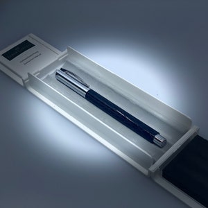 Op de afbeelding: Een donkerblauwe en zilveren pen in een witte etui. De pen heeft een patroon op de schacht en een zilveren clip en punt. De etui bevat een boekje met het Faber-Castell logo.