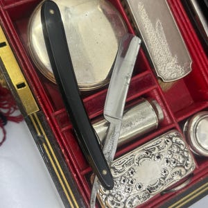 May include: An antique grooming kit with a black handled straight razor, silver-toned compact, and other grooming tools. The kit is housed in a red-lined, black and gold trimmed case, suggesting a vintage aesthetic.