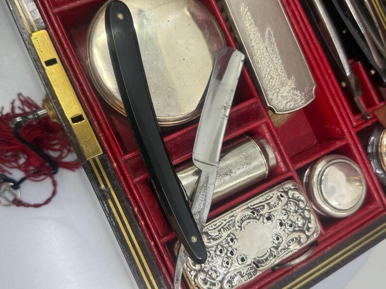 May include: An antique grooming kit with a black handled straight razor, silver-toned compact, and other grooming tools. The kit is housed in a red-lined, black and gold trimmed case, suggesting a vintage aesthetic.