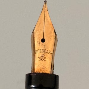 May include: A gold-colored fountain pen nib featuring the engraved text MONTEGRAPPA and the number 585. The nib has a central breather hole and a vertical slit extending to the tip, attached to a black cylindrical grip section.