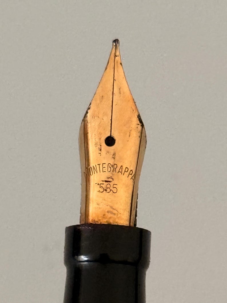 May include: A gold-colored fountain pen nib featuring the engraved text MONTEGRAPPA and the number 585. The nib has a central breather hole and a vertical slit extending to the tip, attached to a black cylindrical grip section.
